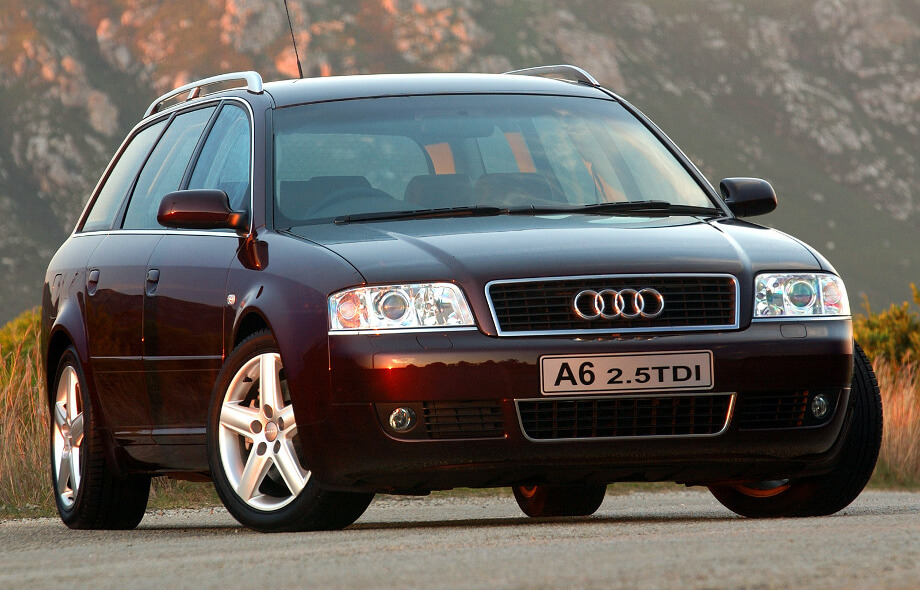На фотографии автомобиль Audi A6 C4. Машина темно-красного цвета, автомобиль стоит на обочине, поросшей травой, рядом красивые горы.
