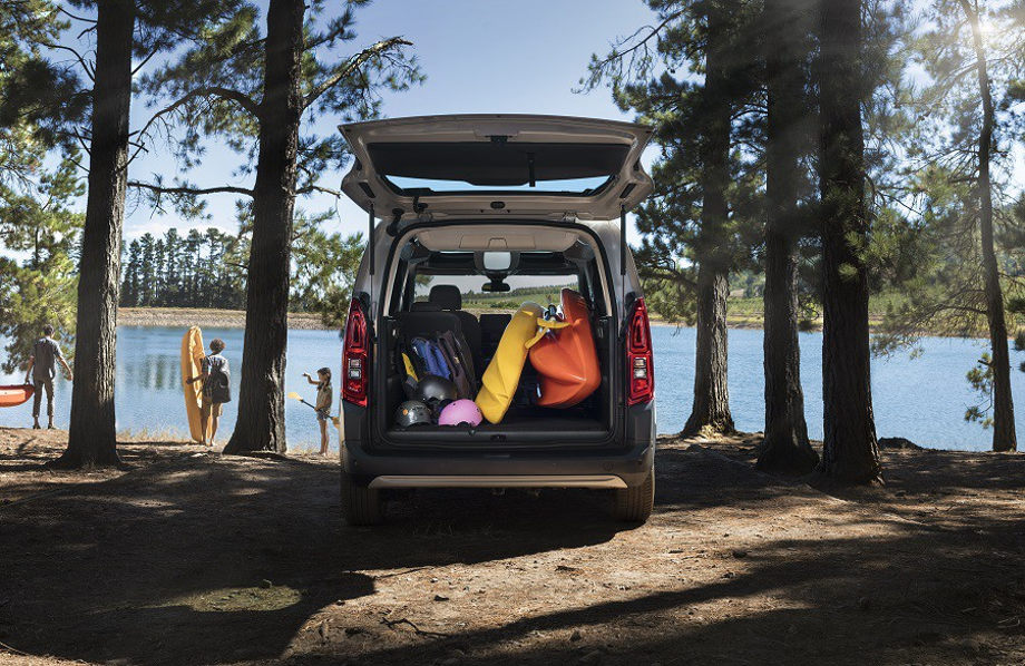 На фото открытый багажник автомобиля Citroen Berlingo III заставлен вещами. На фоне семья отдыхает у реки