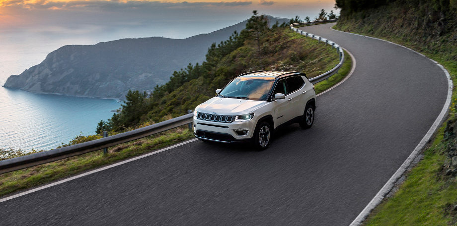 На фото автомобиль Jeep Compass с 2017 белого цвета едет по дороге в горной местности