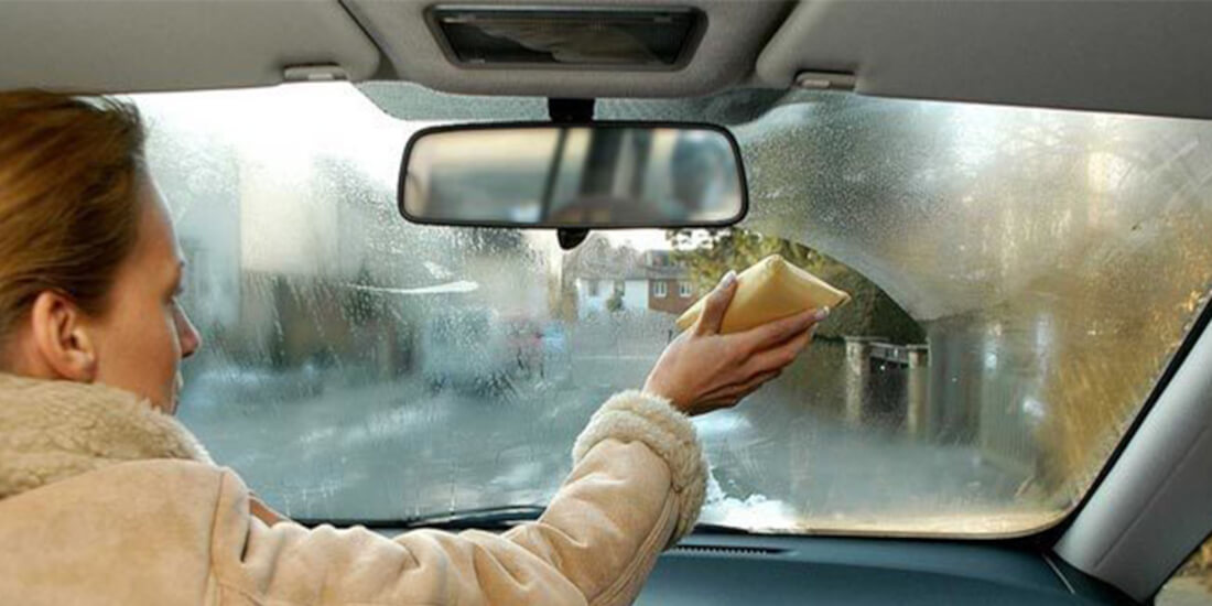 На фото женщина-водитель протирает губкой лобовое стекло изнутри автомобиля, чтобы потом нанести средство против конденсата