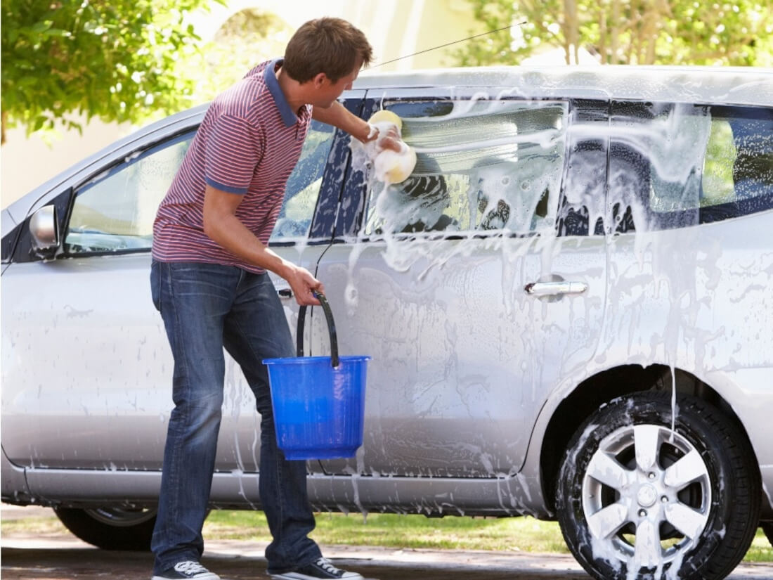На фото чоловік автомобіліст миє машину в домашніх умовах. Він виконує губкою прямі рухи уздовж автомобіля