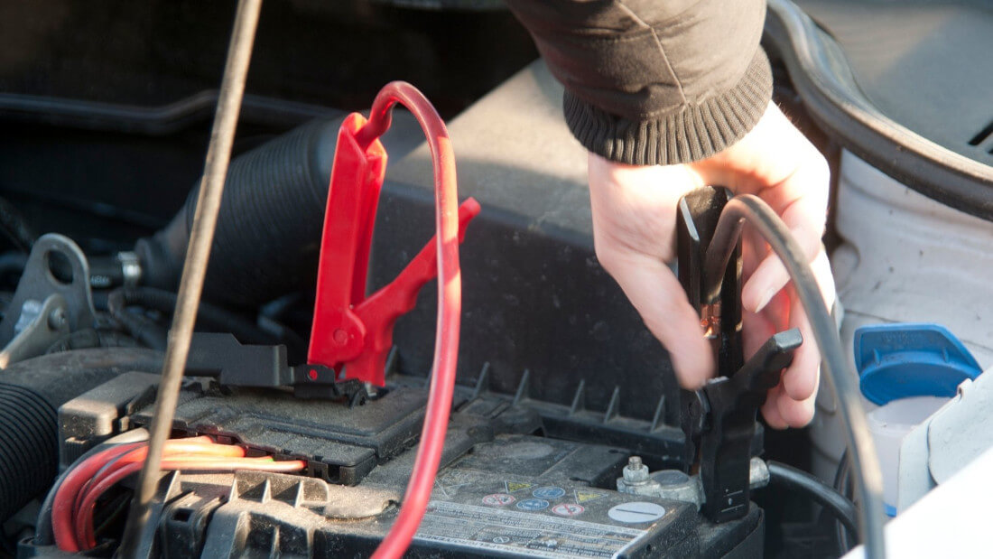 На фото водій підєднує шнури для підзарядки АКБ червоного та чорного кольору до автомобільного акумулятора, який міститься під капотом машини