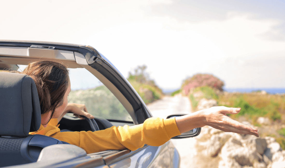 На фото женщина за рулем автомобиля с откидным верхом едет на небольшой скорости и наслаждается поездкой
