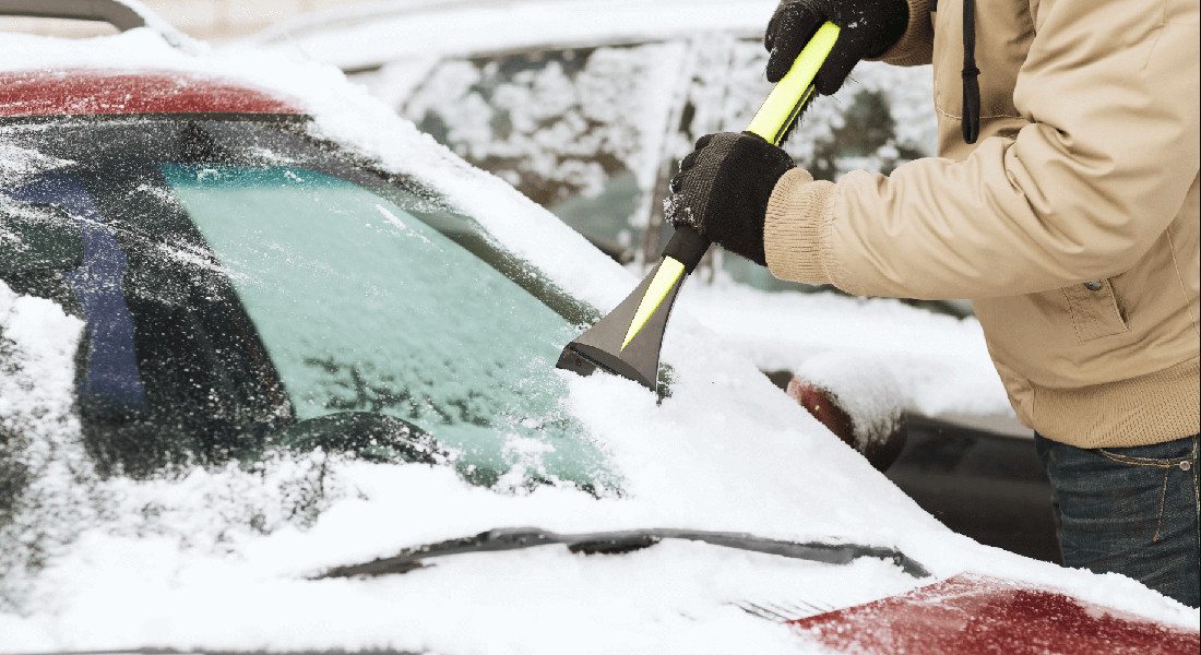 На фото машина присыпана снегом, автомобилист при помощи щетки и скребка очищает лобовое стекло автомобиля от льда