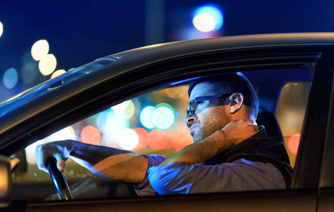 На фото ночью водитель сидит за рулем автомобиля, левой рукой разминает болящую шею, а правой - держится за руль